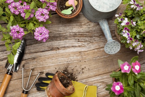 A top-down view of a wooden garden table with various gardening tools and flower pots. The table surface shows natural wood grain and weathering. There are pink and purple flowering plants in terracotta pots along with a small clay pot, some soil, and garden accessories. A grey watering can with a long spout is positioned near the top right, and a pair of hand garden tools with black handles are on the left side. Additionally, a yellow gardening glove and a small garden fork are visible, suggesting ongoing gardening work in a landscaped backyard or outdoor space typical of a garden in the Haringey area. The arrangement highlights the preparation and maintenance of outdoor plantings, supporting gardening services offered by Landscapers Haringey in the local outdoor environment.
