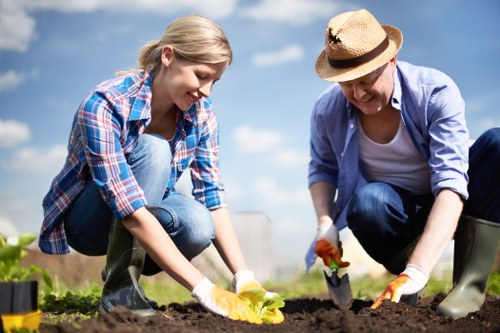 A young woman and an older woman are engaged in gardening activity outdoors in a spacious garden or backyard with a clear blue sky and scattered clouds. The young woman, dressed in a blue and red plaid shirt and jeans, is kneeling on the soil, using her hands to plant a yellow seedling or flower in the freshly turned earth. The older woman, wearing a light blue shirt, dark jeans, a straw hat, and gardening gloves, is crouched beside her, holding a small gardening tool, possibly for digging or planting. The garden features a rich, dark soil bed with well-defined borders and patches of green grass in the background, along with a few garden tools visible nearby. The natural lighting indicates a bright day, contributing to the vibrant colours and clarity of the scene, which illustrates typical outdoor gardening tasks suitable for professional landscaping or lawn care services in the Haringey area, providing a visual representation of gardening efforts and outdoor maintenance activities supported by Landscapers Haringey.