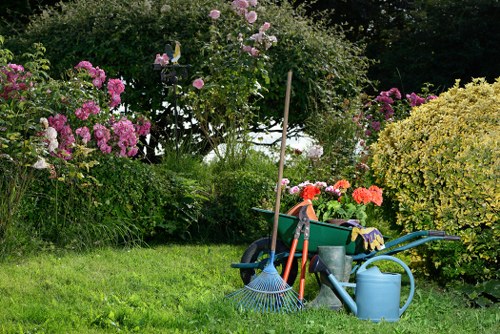The image depicts a well-maintained garden with a lush, green lawn in the foreground, surrounded by various flowering shrubs and bushes, including pink and purple blossoms, as well as yellow-green foliage. A wooden garden trellis supports a flowering rose bush with light pink blooms situated near the center of the scene. In the background, taller trees provide a natural backdrop, and the garden appears to be part of a landscaped outdoor space typical of a residential garden in Haringey. Visible gardening tools and equipment, such as a garden rake, watering can, and a wheelbarrow filled with additional gardening supplies, are arranged on the grass, indicating ongoing maintenance activities. The area benefits from natural daylight, suggesting clear weather, and the scene overall emphasizes an organized and cared-for garden, suitable for professional landscaping or gardening services by Landscapers Haringey.