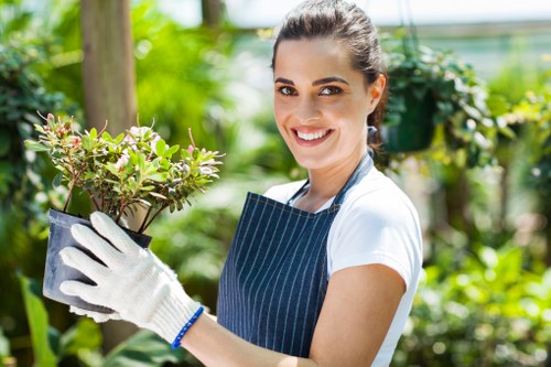 A smiling woman wearing a striped apron and white gloves holds a small potted shrub with green leaves and pinkish flowers in a lush garden setting. The garden features a well-maintained lawn with dense, vibrant greenery, including various bushes and trees, and a mix of natural soil and paved pathways. The background shows taller trees and foliage providing shade, with sunlight filtering through the leaves, indicating a clear day. The scene suggests an outdoor gardening activity, with a focus on plant care and landscape maintenance typical of services offered by companies like Landscapers Haringey, based near the London area.