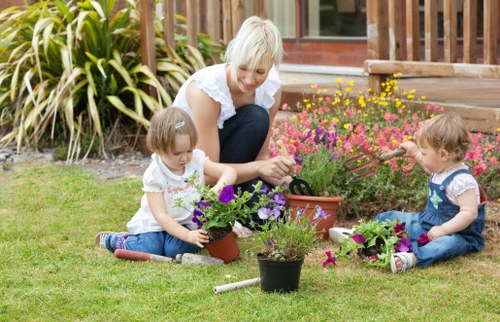 A woman with short blonde hair is sitting on a well-maintained lawn in a garden, engaging with two young children who are planting flowers in pots. The woman is smiling and wearing a white top, while the children, a girl and a boy, are dressed in casual summer clothes; the girl wears a white t-shirt and blue jeans, and the boy wears a blue t-shirt and jeans. They are using small gardening tools and working with colorful flowering plants, including purple, pink, and yellow blooms. The garden background features lush green grass, various plants including tall ornamental grass and a floral bed with bright flowers, and a wooden patio or deck area with a glass door leading to the house. The scene suggests a pleasant, sunny day suitable for outdoor gardening and family activities, emphasizing backyard garden care and planting services provided by landscapers Haringey. The image captures a neat, inviting outdoor space with a focus on natural tones and garden maintenance elements, supporting local gardening and landscaping themes in north London postcodes like N4 or N8.