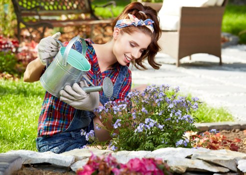 A woman with brown hair tied back wearing a red and blue plaid shirt, grey gardening gloves, and a colourful headband is working in a garden in Haringey, London. She is crouched down next to a flower bed, watering with a metal watering can that has a green lid. The garden features a neatly edged flower bed with blooming purple flowers, bordered by medium-sized grey stones. In the background, there is a paved patio area with outdoor furniture, including a table and chairs, and a lush, well-maintained lawn with a variety of plants and green shrubbery. The scene is illuminated by natural daylight, suggesting a bright, mild day suitable for gardening activities. This setting exemplifies residential gardening tasks such as planting and watering, aligning with professional garden maintenance services offered by Landscapers Haringey without promotional language.