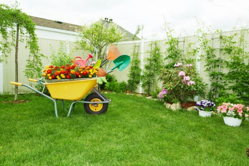 A well-maintained backyard garden in Haringey featuring a lush, green lawn with evenly cut grass in the foreground. The garden is bordered by a white wooden fence, which supports climbing plants with light green foliage running along its length. On the right side, there are colorful potted flowers, including pink, purple, and white blossoms, arranged neatly on the ground near the fence. In the centre of the garden, a small shrub with pink flowers is visible, alongside a larger leafy green bush and a young tree behind it. To the left, a yellow wheelbarrow filled with bright red, yellow, and orange flowers is parked on the grass, with garden tools such as a green watering can and orange rake leaning against it. The scene appears to be outdoors on a cloudy day, with soft natural lighting enhancing the natural tones of the garden. This image showcases elements commonly addressed by professional gardening services like those offered by Landscapers Haringey, including lawn care, plant maintenance, and outdoor space enhancement.