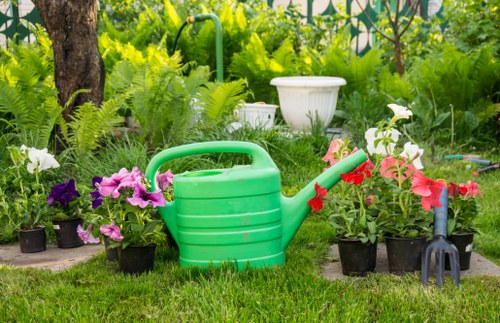 The image depicts a residential garden area with a neatly maintained lawn, surrounded by an array of flowering plants and potted flowers in shades of purple, white, and pink. In the foreground, a green plastic watering can is placed on the grass next to a collection of potted plants, including some with large, colorful blossoms. To the right, a small garden fork is partially visible, positioned amongst the flower pots. The background features taller greenery, such as shrubs and leafy plants, and a garden tree with a sturdy trunk, contributing to a lush, vibrant outdoor environment typical of a well-maintained garden in Haringey. The overall scene suggests attention to outdoor gardening maintenance, with natural daylight illuminating the space, and no human activity present. The setting aligns with residential gardening and landscaping services provided by companies like Landscapers Haringey, emphasizing garden care elements including planting, watering, and tidying outdoor spaces in the local area.