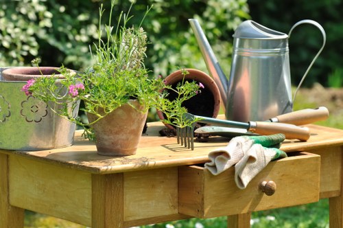 A close-up view of a wooden garden table in an outdoor setting, featuring several gardening tools and accessories including a silver watering can, a pair of gardening gloves, a small rake, and terracotta pots with green plants and pink flowers. The background displays a lush, green garden with leafy shrubs and trees, suggesting a well-maintained outdoor space suitable for gardening and landscaping services. The sunlight creates a bright and inviting atmosphere, highlighting the natural tones of the wood, soil, and foliage, with the scene reflecting an active gardening environment consistent with services offered by Landscapers Haringey in the Haringey area.