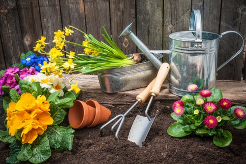 The image shows a section of a landscaped garden with a focus on gardening tools and flower arrangements. In the foreground, there is freshly turned dark soil, indicating recent planting activity. To the left, a vibrant cluster of potted primroses in yellow, purple, and white colors adds a splash of natural colour, while on the right, a pot of pink and yellow primulas is visible. Behind these, various gardening tools lie on the soil, including a hand rake, a trowel, and a small fork, all with wooden or metal handles. A weathered metal watering can is positioned towards the back, next to a small plant watering container. An empty terracotta pot is placed upside down, suggesting ongoing planting or container gardening. The background features a wooden fence, which provides a natural backdrop for the landscaped outdoor space, creating a neat and functional garden area suitable for flower bed creation and general outdoor maintenance by local gardening professionals such as Landscapers Haringey, serving the Haringey postcode area in North London. The overall scene is set in natural daylight, with no harsh shadows, illustrating an organized and inviting garden space ready for further planting or landscaping work.