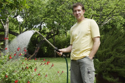 A man with light brown hair, wearing a pale yellow T-shirt and grey cargo shorts, stands in a well-maintained garden in Haringey, London, holding a garden hose and watering a flowering shrub with small red blooms. The garden features a lush, green lawn with dense trees and bushes in the background, providing shade and natural privacy. The scene captures an outdoor space equipped for gardening and lawn care, with healthy, vibrant plant life and a neatly landscaped environment. The lighting is natural, suggesting a bright, clear day, ideal for outdoor maintenance and gardening activities. This setting exemplifies a typical London garden that could benefit from professional gardening services to enhance its appearance, aligned with the offerings of companies like Landscapers Haringey, who support local outdoor maintenance and landscaping needs.
