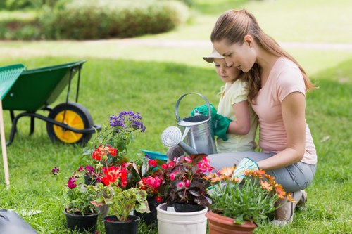 A woman and a young girl are working together in a well-maintained backyard garden, tending to an array of vibrant flowering plants in pots, including petunias and geraniums, which display shades of red, pink, and purple. The garden features a lush, green lawn with neatly edged flower beds and a mix of blooming shrubs. In the background, there are taller trees providing shade and a garden wheelbarrow with yellow wheels positioned on the grass, indicating ongoing outdoor maintenance. The scene takes place on a bright, sunny day with natural light illuminating the outdoor space, suggesting a pleasant environment suitable for gardening activities. The image conveys a focus on gardening tasks such as potting and plant care, reflecting professional landscaping and garden maintenance services typical of a garden in Haringey, North London, with an emphasis on creating welcoming outdoor spaces for families and gardening enthusiasts through careful planting and lawn upkeep by companies like Landscapers Haringey.
