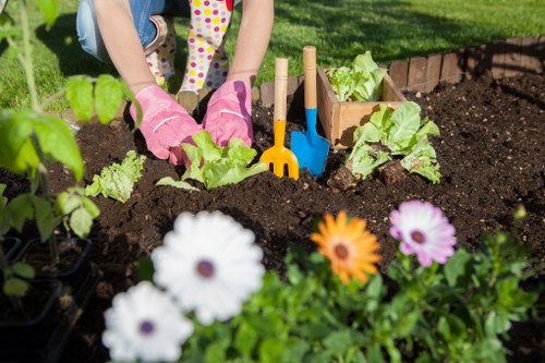 The image shows a person gardening in a residential backyard, engaged in planting young leafy green vegetables, possibly lettuce or similar, in dark, rich soil. The gardener's hands are protected with pink gardening gloves, and they are surrounded by small gardening tools with yellow and blue handles, including a trowel and a hand rake, placed in the soil near the plants. In the foreground, there are vibrant flowering plants with white, orange, and purple blossoms, providing a colourful contrast to the leafy greens. The garden features a well-maintained lawn in the background with neatly edged flower beds, bordered by a layer of bricks or stones, indicating careful landscaping and outdoor maintenance typical of UK gardens. The scene is illuminated by natural daylight under clear weather conditions, reflecting a tidy, productive outdoor space, and highlighting the values of gardening and planting services offered by companies like Landscapers Haringey in the N22 postcode area.
