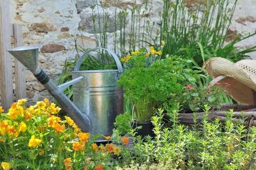 The image depicts a small garden area with a variety of plants and garden tools arranged against a rustic stone wall. In the foreground, there are bright yellow and orange flowering plants, possibly marigolds, with dense green foliage surrounding them. Behind these flowers, a silver metal watering can is placed, alongside a weathered straw hat resting nearby. To the right of the watering can, there is a lush, leafy green plant, possibly a herb, in a container. Taller grass-like plants and other greenery grow along the stone wall in the background, providing a natural backdrop. The garden appears well-maintained, with a mix of flowering and foliage plants, and the environment suggests a casual, outdoor gardening space suitable for planting, watering, and general garden care, aligning with landscaping services offered by Landscapers Haringey in the Haringey area. The scene is under natural daylight, indicating a clear, possibly warm day, with vibrant colours and textures emphasizing an inviting outdoor setting for gardening and outdoor maintenance tasks.