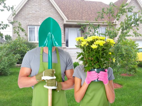 In a suburban garden setting, two individuals are preparing for gardening activities; one person, wearing a teal gardening helmet, grey shirt, green gloves, and a green apron, is holding a large garden spade with both hands, while the other person, wearing a grey shirt and pink gloves, is holding a vibrant bouquet of yellow flowers in front of their face, obscuring their identity. The garden features a well-maintained green lawn in the foreground, bordered by shrubbery and small plants, with a paved pathway leading towards a residential house with light brick walls and white-framed windows in the background. The scene is brightly lit, suggesting a clear day, with natural light illuminating the lush foliage and the fresh flowers, highlighting the outdoor space's nurturing environment which is characteristic of professional gardening and landscaping services offered by Landscapers Haringey in the Haringey area, close to the N22 postcode, creating an inviting and orderly outdoor space.