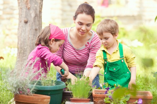 A woman with dark hair tied back, wearing a pink and white striped long-sleeve top, is in a garden with two young children; a girl in a pink jacket and a boy dressed in a yellow shirt with green overalls. They are engaging in gardening activities around a table with various potted plants, including herbs, flowers, and foliage. The garden features a background of trees, with a lawn area in the foreground covered with lush, green grass and flower beds along a natural stone border. The scene is illuminated by natural daylight, suggesting a mild weather day, and the setting reflects a well-maintained outdoor space suitable for gardening and family outdoor activities, which aligns with the services offered by Landscapers Haringey in the local area of Haringey postcode, fostering a natural connection to gardening and outdoor maintenance services.